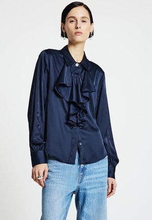 Frau trägt eine marineblaue Satinbluse mit Rüschen und einem dekorativen Knopf, kombiniert mit hellblauen hoch taillierten Jeans vor weißem Hintergrund.