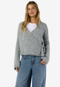 Ung kvinna med en grå fluffig wrap-tröja över en vit topp och lösa blå jeans, stående med en hand i fickan.