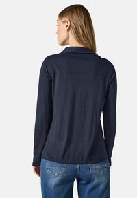 Camicia a maniche lunghe blu navale con colletto, texture morbida, dettaglio cucitura sullo yoke posteriore, abbinata a jeans azzurri chiari. Design casual discreto.