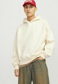 Cremefarvet hoodie med et hyggeligt design, tætsiddende manchetter og en blød tekstur. Kombinationen er med løstsiddende blå denimbukser og en rød kasket.