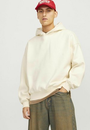Hoodie crème au design douillet, avec des poignets ajustés et une texture douce. Associé à un pantalon en denim bleu ample et une casquette rouge.