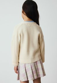 Pull en tricot côtelé crème avec épaules tombantes, associé à une jupe plissée rose pastel présentant un motif écossais aux lignes douces bleu et vert.