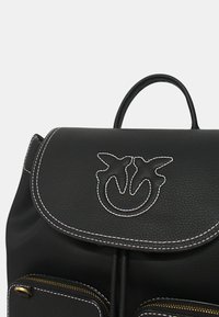 Zaino in pelle nera con un pattina arrotondata, cuciture bianche, un logo decorativo e due tasche frontali con cerniera. Presente maniglia superiore.