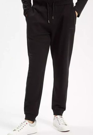 Osoba w czarnych spodniach dresowych typu jogger z elastycznymi ściągaczami i sznurkiem w pasie, w połączeniu z białymi niskimi trampkami na białym tle.