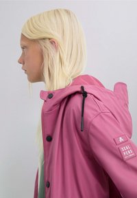 Chaqueta impermeable rosa con capucha, botones negros y un parche en la manga con un logo de calavera. Textura suave y diseño casual.