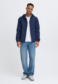 Navy hooded jacket with two front pockets, paired with light blue jeans and gray sneakers. Smooth fabric with a relaxed fit.