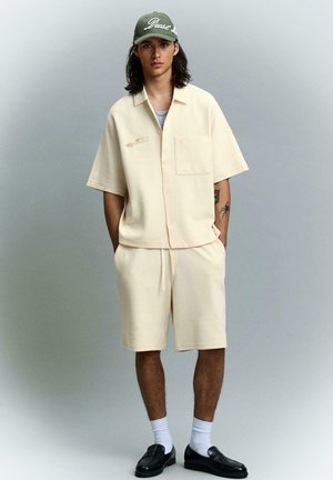 Mladý muž s dlhými vlasmi, ktorý nosí béžovú krátkym rukávom košeľu a šortky, biele ponožky, čierne mokasíny a zelenú šiltovku, stojí s rukami v kapsách.