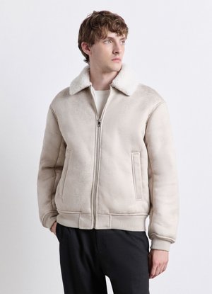 Young man wearing beige shearling jacket with white fur collar and black pants, looking to the side against plain background.