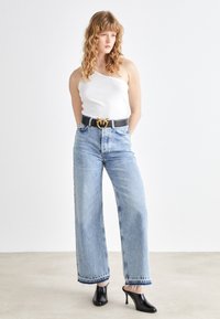 Light blue wide-leg jeans with frayed hems, paired with a white one-shoulder top and a black belt featuring a gold emblem. Black heels.