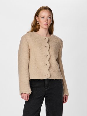 Vaalean beige, teksturoitu neulepaita, jossa on aallotettu helma ja viisi koristeellista metallipainiketta. Paidassa on pitkät hihat ja pyöreä kaula-aukko.