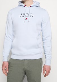 Sudadera azul claro de tela suave con un bolsillo tipo canguro en la parte frontal. Presenta el logo "TOMMY HILFIGER" en negro y detalles de rayas roja y blanca en la capucha.