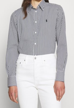 Femme portant une chemise boutonnée à manches longues avec des rayures verticales noires et blanches, rentrée dans un pantalon blanc taille haute.