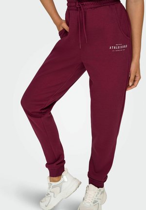 Burgundie Jogginghose aus weichem Baumwollmaterial, mit seitlichen Taschen, elastischem Bund und Rippenbündchen, mit einem weißen Logo auf der Vorderseite.