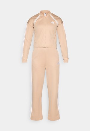 Beżowy dres uszyty z miękkiego materiału, posiadający zapinaną na zamek kurtkę z wysokim kołnierzem oraz białe paski wzdłuż rękawów i nogawek. Dyskretny detal z logo.