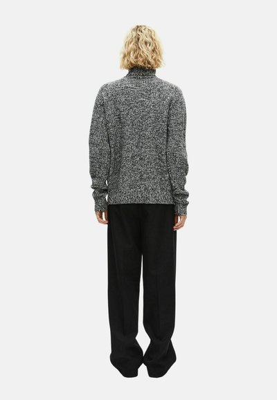 Asmuo su trumpais garbanotais šviesiais plaukais, vilkintis pilką megztinį ir laisvas juodas kelnes, stovi atsukęs nugarą baltame fone.