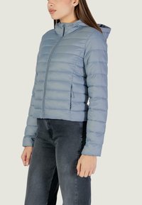 Giacca imbottita azzurro chiaro con motivo trapuntato orizzontale, chiusura con zip e cappuccio. Tessuto strutturato e design aderente, abbinata a pantaloni grigi.