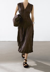 Brown, sleeveless dress with vertical pleats, knee-length, paired with a matching bag and chunky black sandals with ankle straps.