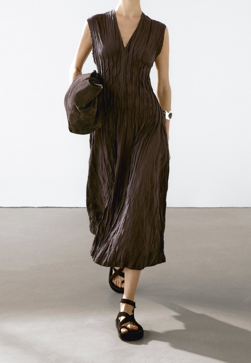 Brown, sleeveless dress with vertical pleats, knee-length, paired with a matching bag and chunky black sandals with ankle straps.
