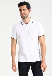 Polo bianco con rifiniture del colletto rosse e blu navy, maniche corte e un logo discreto sul petto. Abbinato a jeans scuri.