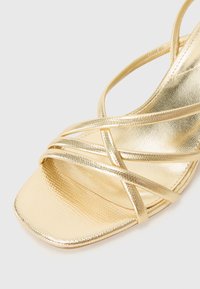 Gold strappy sandal with a textured metallic surface, square toe, and multiple crisscrossed straps for secure fit. Flat sole design.