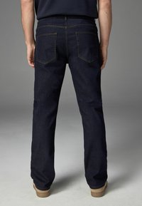 Dunkelblaue Denim-Jeans mit geradem Bein, ausgestattet mit zwei Gesäßtaschen und braunen Nähten, kombiniert mit hellbraunen Schuhen.