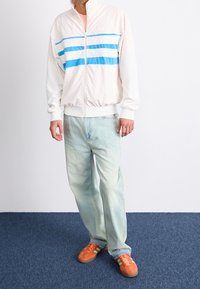 Man i vit zip-up jacka med blå ränder, ljus tvättade jeans och orangea sneakers som står på en blå matta mot en vit bakgrund.