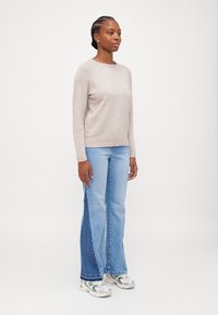 Pull en tricot beige avec un col rond, associé à un jean évasé bleu clair avec un panneau en denim plus foncé contrastant. Baskets blanches.