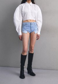 Persoon in een witte korte lange mouw shirt, lichtblauwe high-waisted denim shorts met uitsnijdingen, en zwarte kniehoge laarzen.