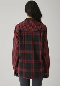 Femme aux longs cheveux bruns, portant une chemise en flanelle à carreaux rouges et noirs, vue de dos, se tenant devant un fond clair.