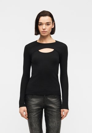 Frau mit kurzen dunklen Haaren, die ein schwarzes Langarmoberteil mit Ausschnitt am Oberkörper und strukturierte schwarze Lederhosen trägt, steht aufrecht.