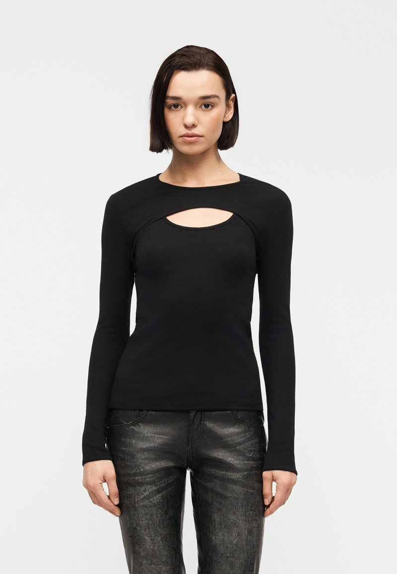 Γυναίκα με κοντά σκούρα μαλλιά, φορώντας μαύρο μακρυμάνικο τοπ με άνοιγμα στο στήθος και μαύρο δερμάτινο παντελόνι με υφή, όρθια.