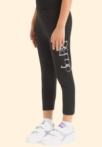 Leggings neri con un design aderente e grafica bianca sul lato, abbinati a sneakers bianche con dettagli viola.
