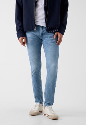 Mann trägt hellblaue Slim-Jeans, weiße Sneaker, weißes T-Shirt und marineblaue Zip-Jacke, steht vor einem einfarbigen Hintergrund.