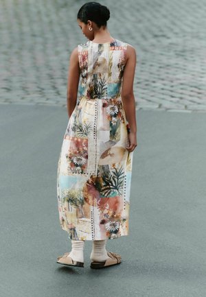 Femeie cu părul închis la culoare prins în coc, purtând o rochie colorată cu patchwork floral, mocasini bej și șosete albe, stând pe o suprafață pavată.