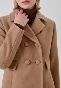 Kamelfarbener Wollmantel mit zweireihigem Design, großen runden Knöpfen und einem Reverskragen, der eine strukturierte Oberfläche und Seitentaschen aufweist.