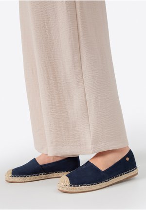 Füße mit marineblauen Espadrilles mit gewebten Jutesohlen, kombiniert mit beigen, weit geschnittenen, strukturierten Hosen.