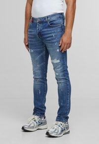 Moški v ozkih, raztrganih modrih kavbojkah in srebrnih športnih copatih, stoji ob enobarvni svetli podlagi.