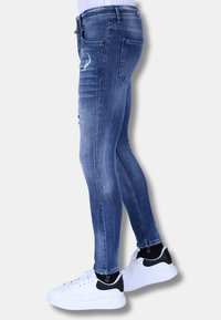 Slanke blauwe jeans met een vervaagde wassing, met gescheurde elementen en subtiele whiskering. Combinatie met witte sneakers en zwarte accenten.