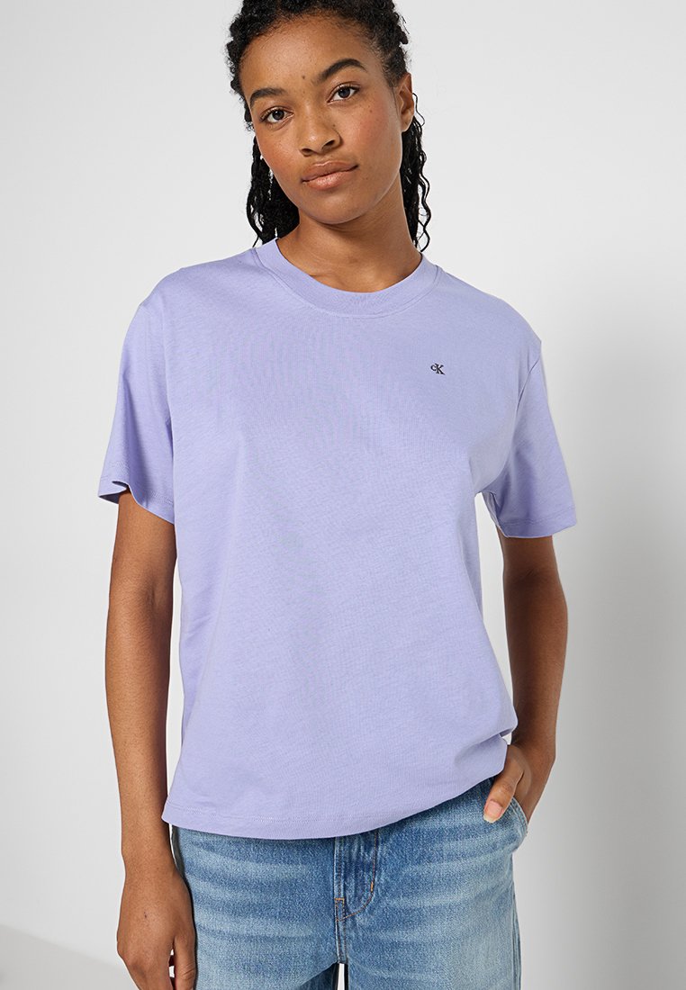 Młoda osoba z kręconymi włosami, ubrana w jasnofioletową koszulkę CK i niebieskie dżinsy, ręka swobodnie w kieszeni, neutralne tło.