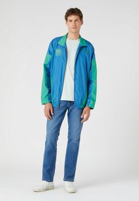 Blue and green windbreaker jacket with elastic cuffs and hem, zip-up front, adorned with logo print. Worn with light green shirt and denim jeans.