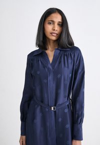 Abito blu navy a maniche lunghe realizzato in tessuto morbido, con un sottile design a motivo, colletto e una vita cintrata con una cintura a fibbia argentata.