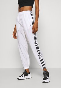 Pantalon de sport blanc avec des bandes latérales noires, taille élastique et logos Adidas à l'avant. Associé à des baskets noires et blanches.