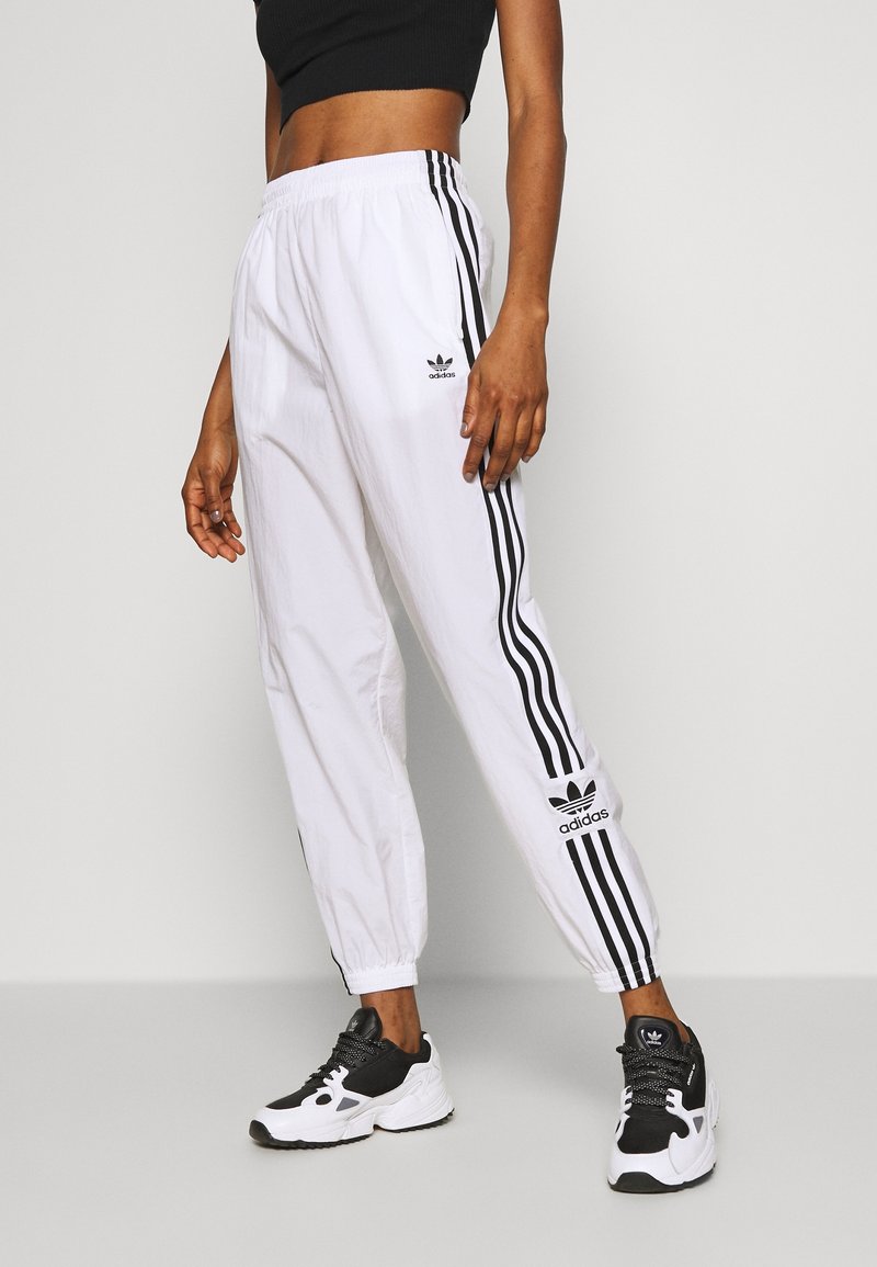 Pantalon de sport blanc avec des bandes latérales noires, taille élastique et logos Adidas à l'avant. Associé à des baskets noires et blanches.