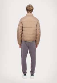 Jachetă puffy de culoare bej, cu guler înalt și cusături orizontale, asociată cu pantaloni joggeri gri. Dispune de manșete elastice și un logo discret pe mânecă.