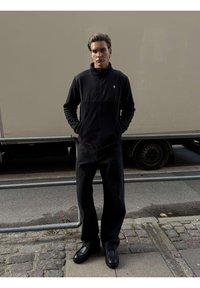 Młody mężczyzna w czarnej kurtce na zamek, czarnych szerokich spodniach i czarnych butach stoi na brukowanej ulicy z rękami w kieszeniach.