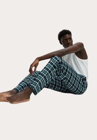 Geruite pyjamabroek in groen, donkerblauw en wit, met een losse pasvorm en zachte stof. Gemaakt met een witte tanktop en blote voeten. Zittende pose.