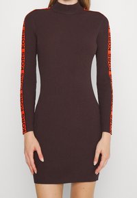 Gros plan sur une robe marron côtelée ajustée à manches longues, ornée de rayures orange vif et du texte "KORS" le long des bras.