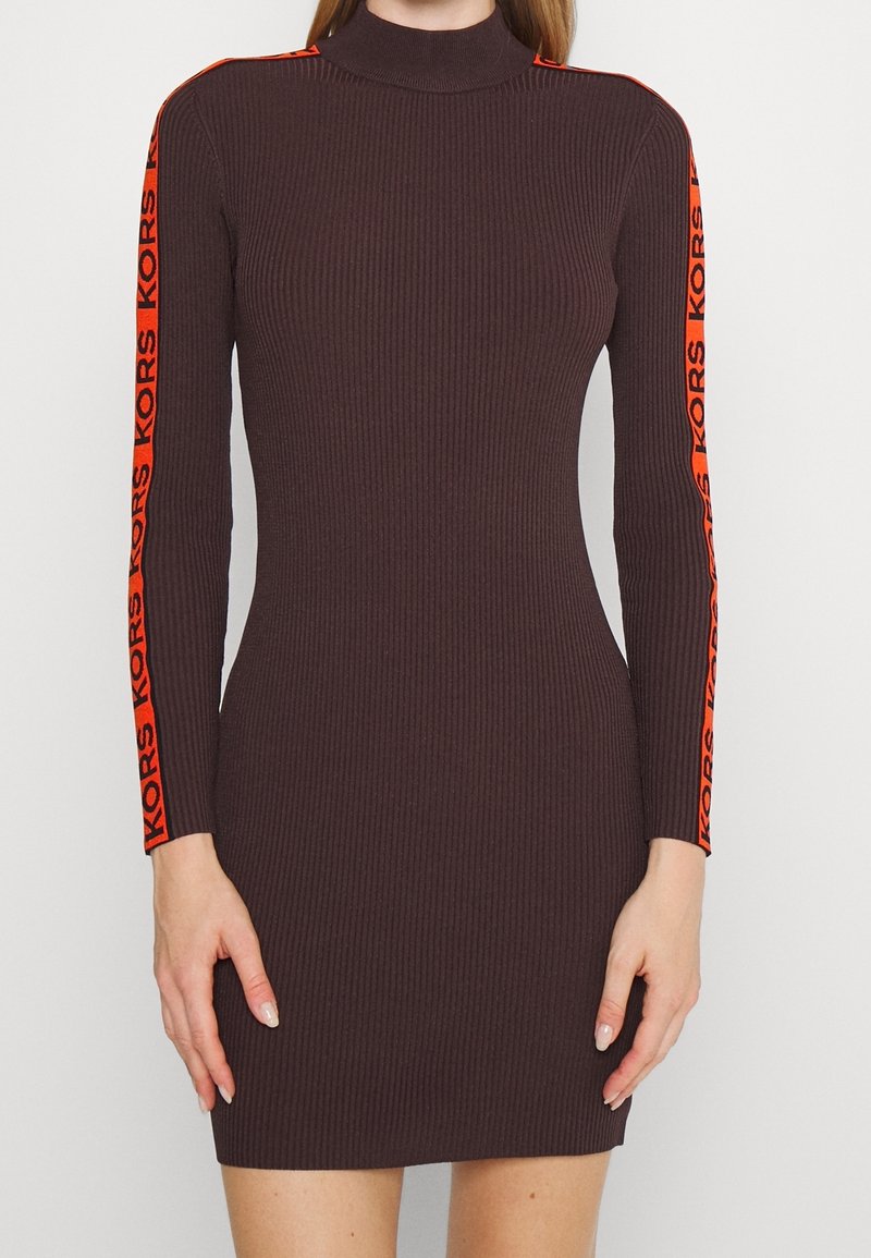 Gros plan sur une robe marron côtelée ajustée à manches longues, ornée de rayures orange vif et du texte "KORS" le long des bras.