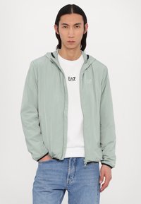 Lys grøn, letvægtsjakke med hætte, lynlås foran og sidelommer. Båret over en hvid sweatshirt, parret med blå jeans.
