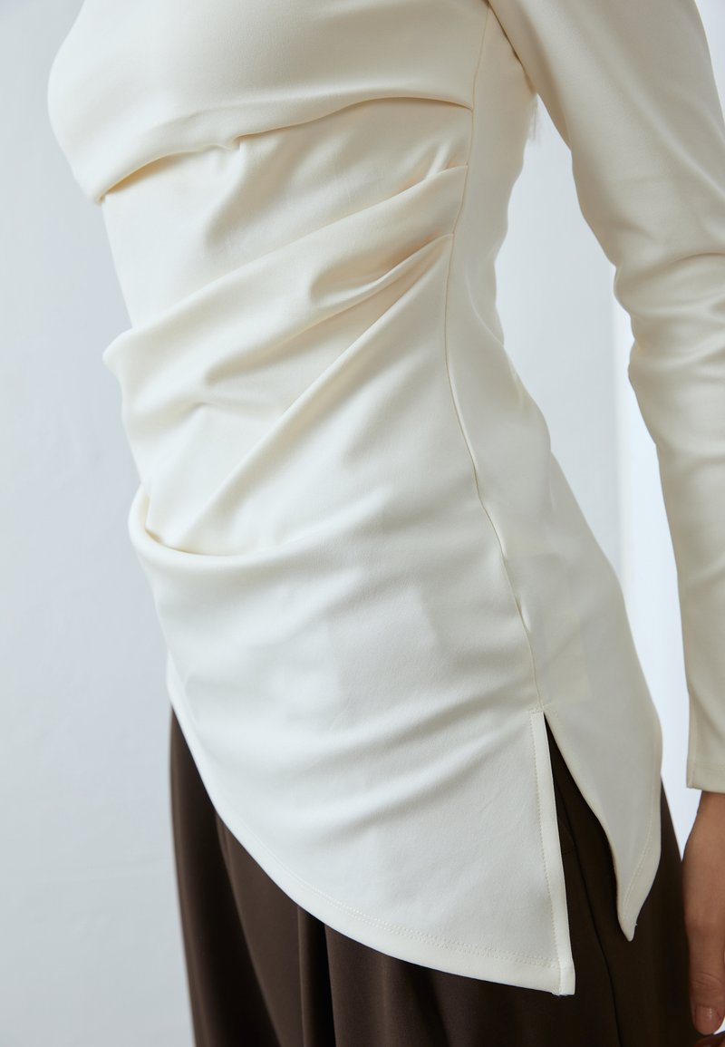 Kremowa bluzka z długim rękawem z marszczeniami i bocznymi rozcięciami, wykonana z miękkiego materiału. Połączona z szerokimi spodniami w ciemnym brązie.